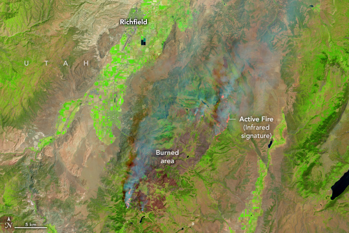A false-color satellite image shows a wildland fire burning in Utah. Smoke billows from areas of active fire along the edges of the burned area (dark brown). The infrared signature from active burning is shown in orange. Areas of unburned vegetation (light green) and dry land (light brown) lie on either side of the fire.
