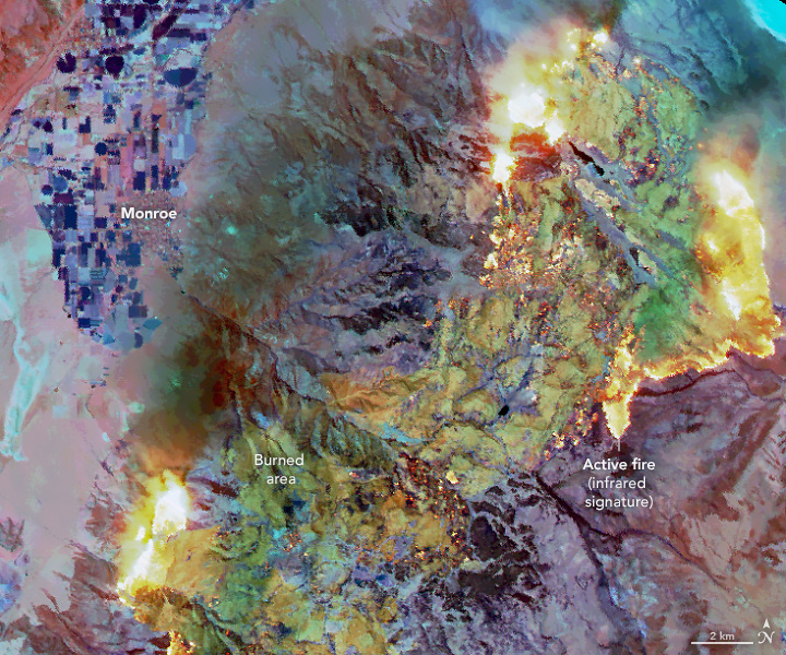 A false-color aerial image shows the same fire later in the day. Actively burning areas (yellow) have grown compared to the earlier image. Active fire, smoldering, and recently burned areas (yellow, red, and light green) contrast with the surroundings (shades of blue and purple).