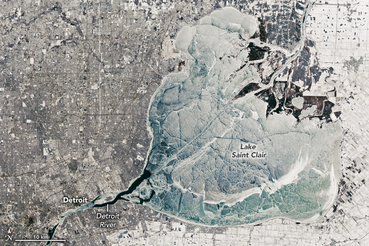 A satellite image shows the heart-shaped Lake Saint Clair covered in blue-to-white ice with large cracks running through it. The surrounding urban and agricultural landscape is snow-covered. On the lake’s northeast side, the top of the heart, a river splays into a delta. The Detroit River flows past the city of Detroit on the opposite side.