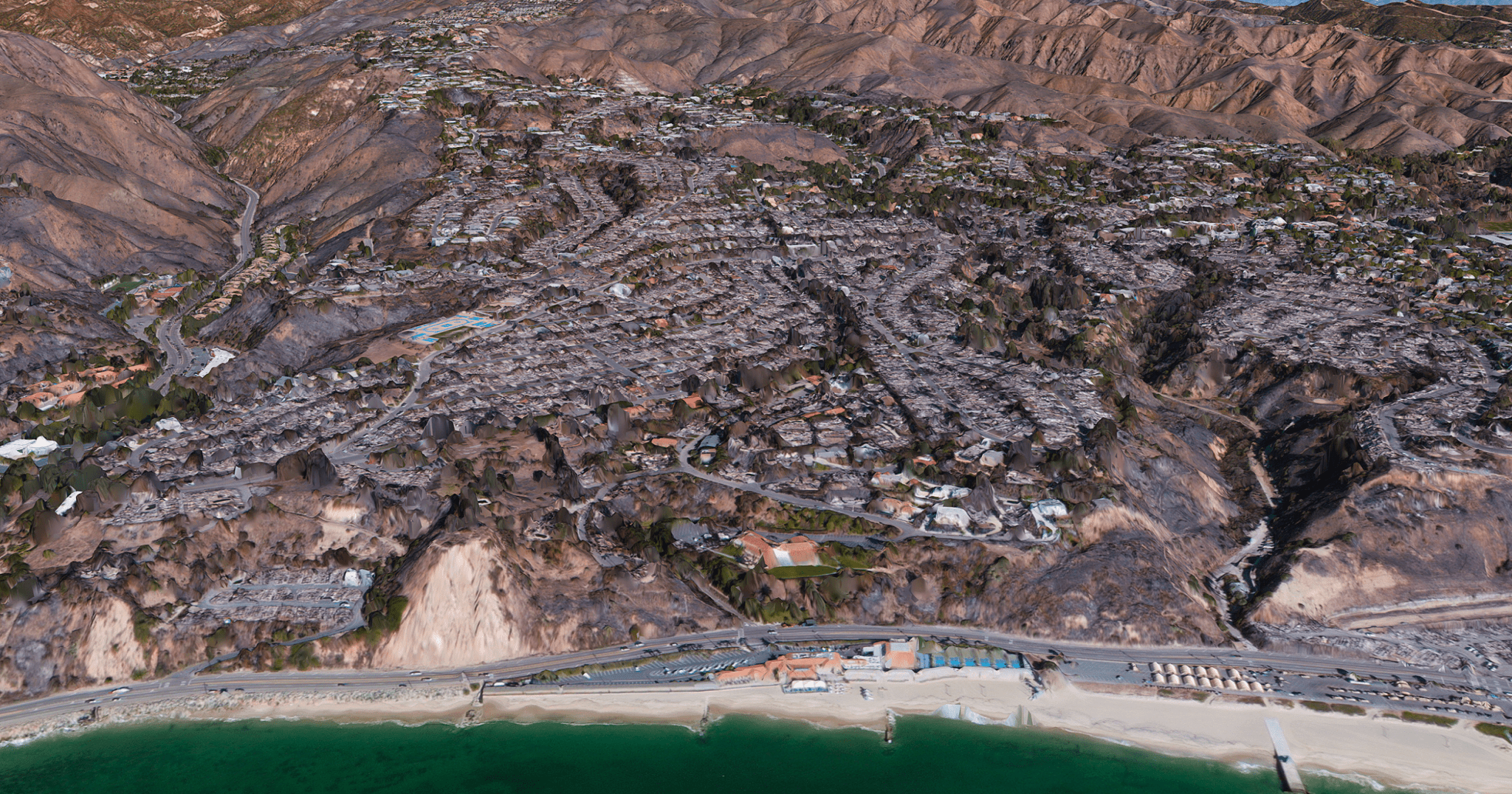 Vantor aerial view of a coastal neighborhood heavily damaged by wildfire, with widespread structural destruction, charred terrain, and the ocean visible in the foreground.