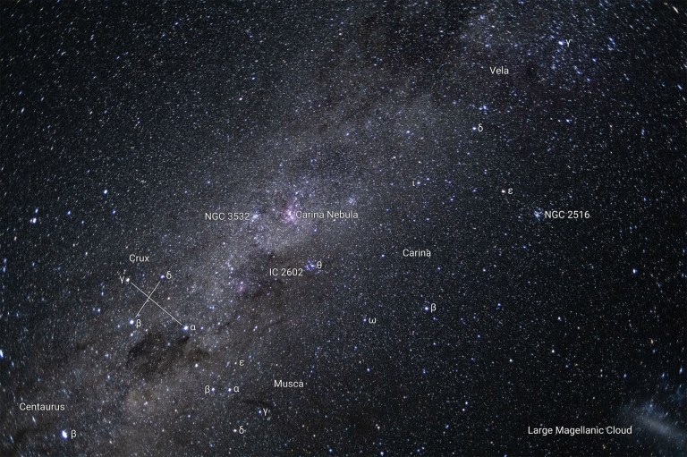 A starfield filled with thousands of stars and shining clouds of dust. The sky is black behind the stars. The Milky Way streaks diagonally across the sky in a white and pink cloud, from the lower left to the upper right.