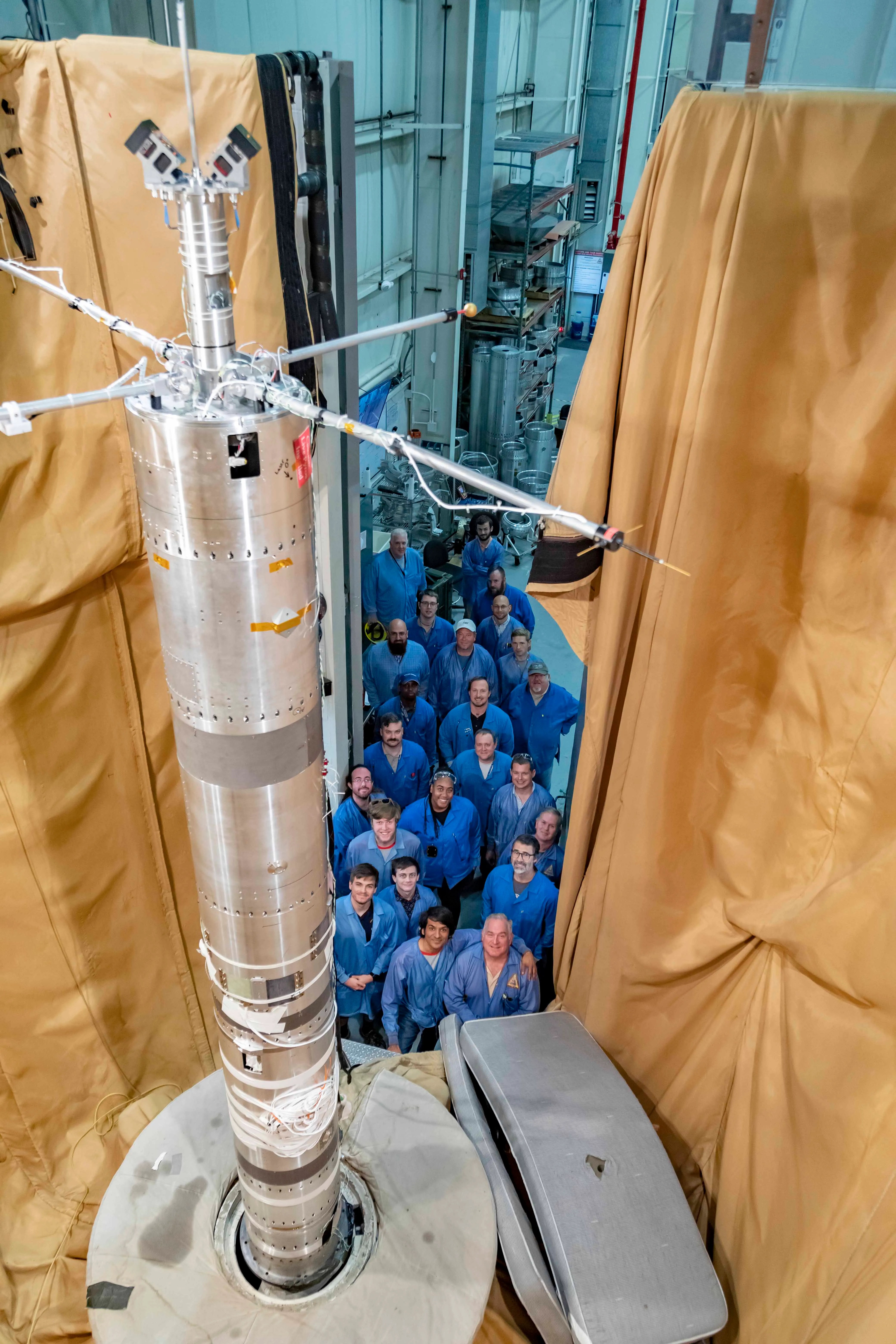 A group of people in blue lab coats stands around a tall, metallic rocket component inside an industrial facility with beige protective curtains.