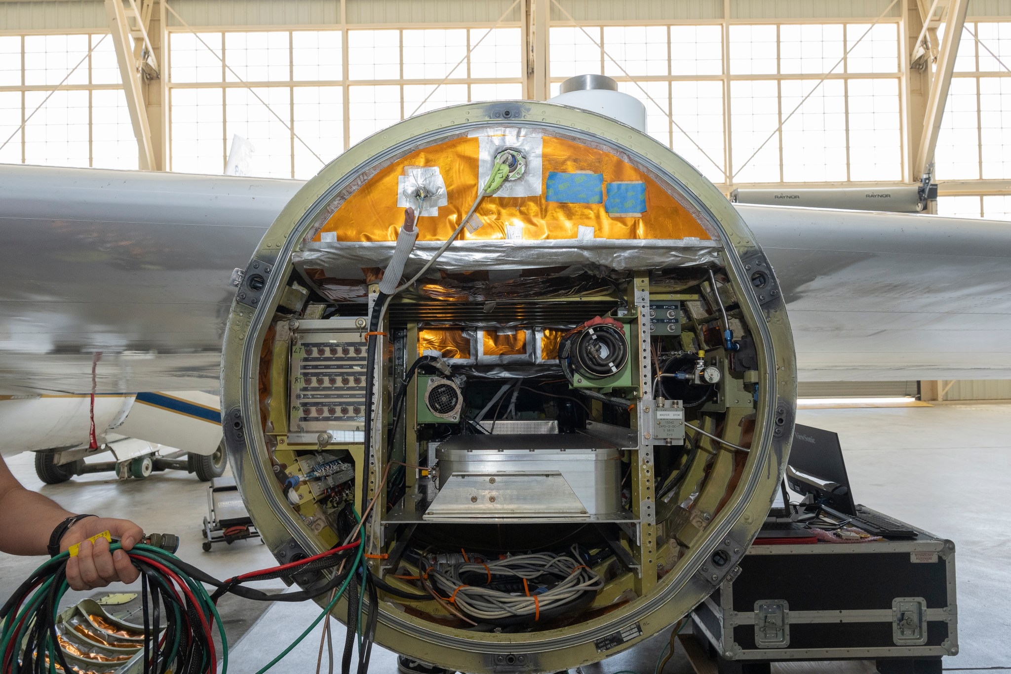 Front view of an open instrument pod mounted beneath a NASA ER‑2 aircraft, showing cables, sensors, and electronic equipment arranged inside a circular housing. A person’s hand holding a bundle of cables is visible on the left side, with the aircraft hangar visible in the background.